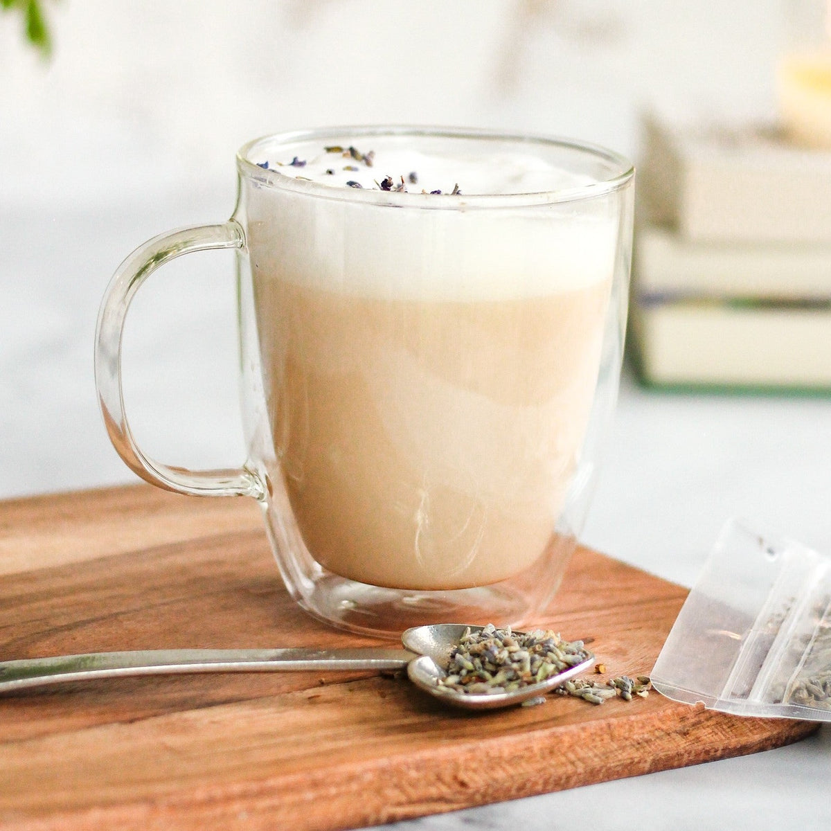 A foamy, lavender-sprinkled latte made with Coco-Lavender Chai in a glass mug, with an Heirloom Heart-Shaped Pewter Spoon full of Coco-Lavender Chai beside it. Both are on top of a wooden cutting board, with a stack of books in the distance.