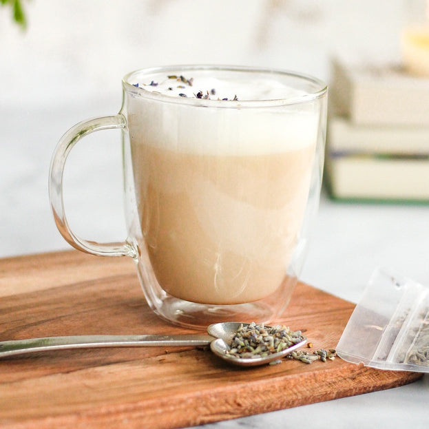 A foamy, lavender-sprinkled latte made with Coco-Lavender Chai in a glass mug, with an Heirloom Heart-Shaped Pewter Spoon full of Coco-Lavender Chai beside it. Both are on top of a wooden cutting board, with a stack of books in the distance.